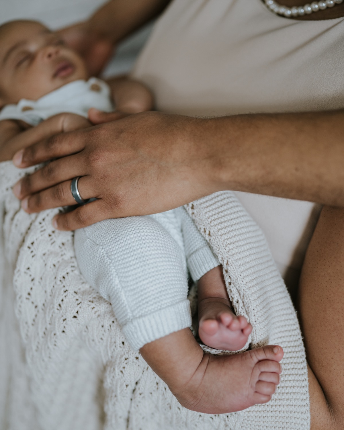 Father's hand with wedding ring resting on sleeping newborn's body, tiny feet peeking out of knit blanket