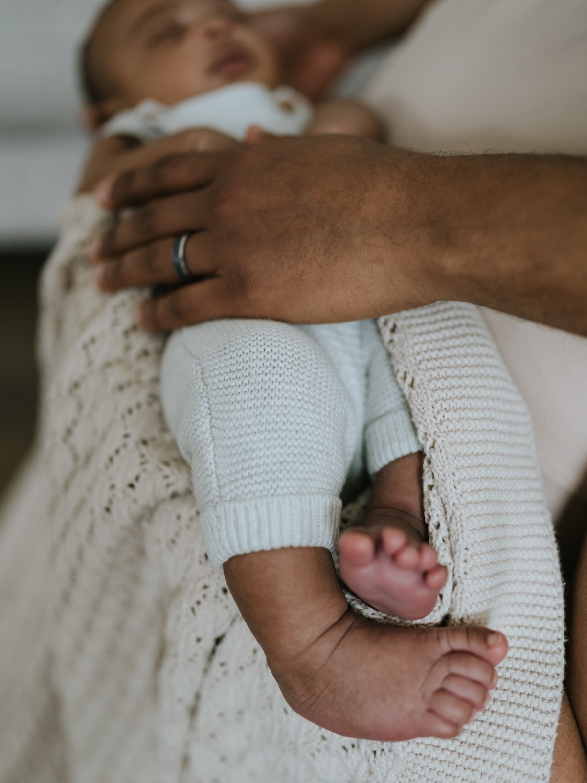 Detail shot of father's hand holding baby, with newborn's small feet and knit outfit in focus