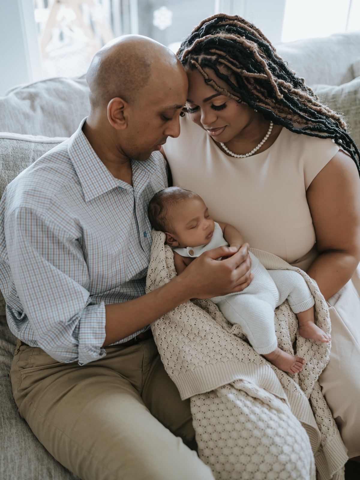 Parents sitting on the couch, foreheads touching, holding newborn Yohannes between them in a knit blanket