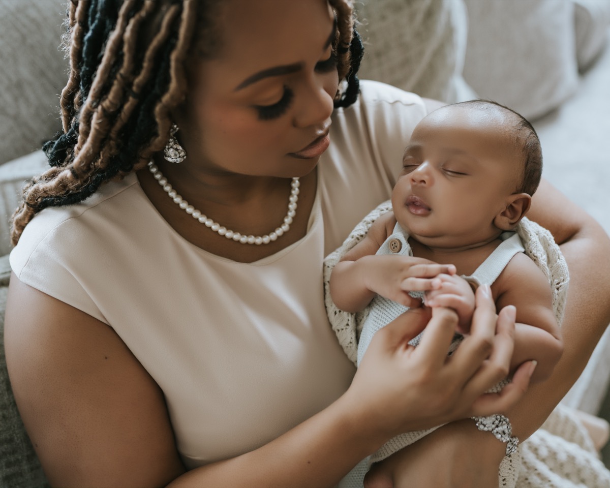 Mother with locs and pearl necklace holding sleeping Yohannes, gently touching his tiny hands